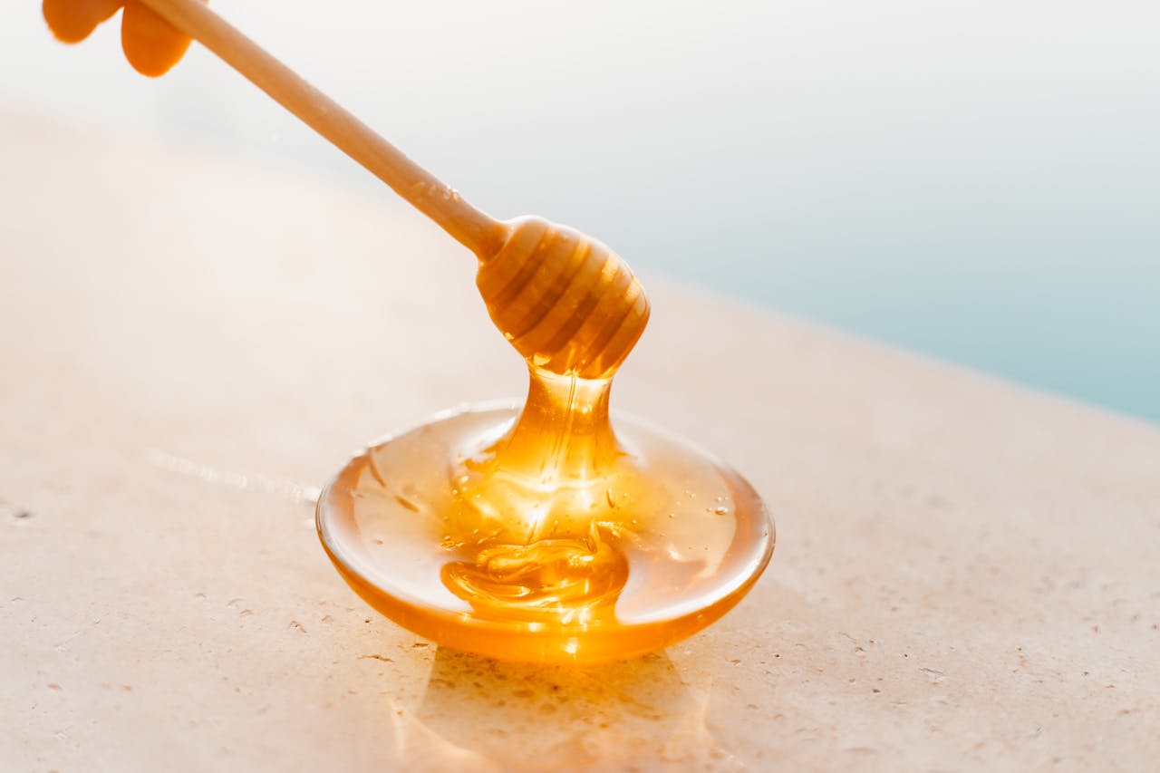home-hero Golden honey flowing from a wooden dipper onto a glass plate, captured in warm daylight.